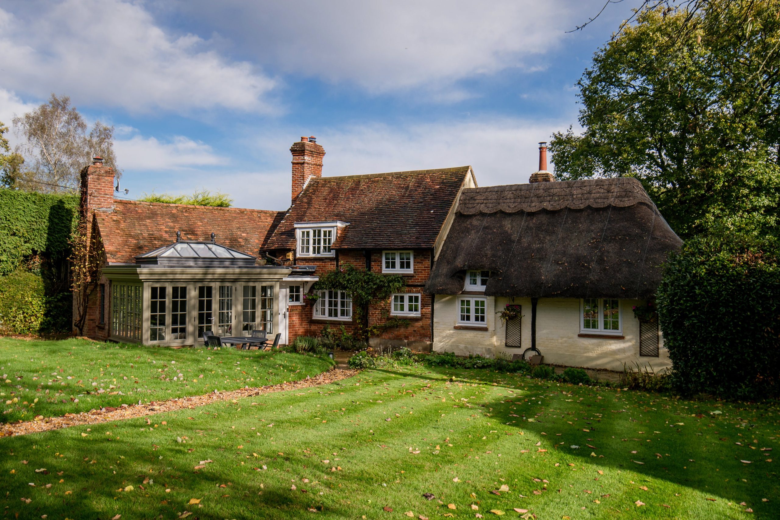 Traditional thatched cottage with white timber windows and a glazed garden extension, set within a landscaped lawn and mature trees under a blue sky.