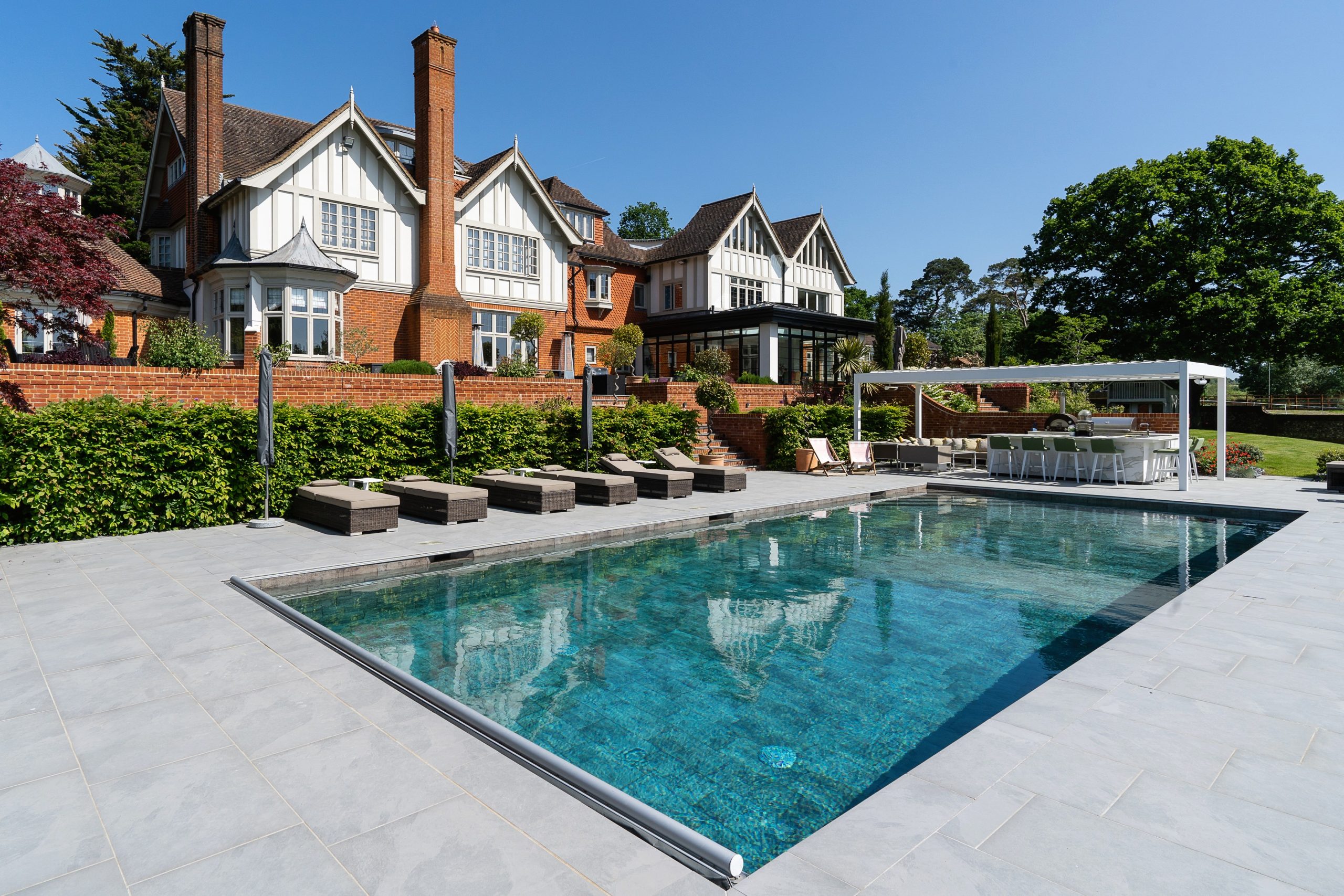 Large traditional detached house with white gables and red brick, overlooking a landscaped swimming pool area with sun loungers, paved terrace, and outdoor dining space.