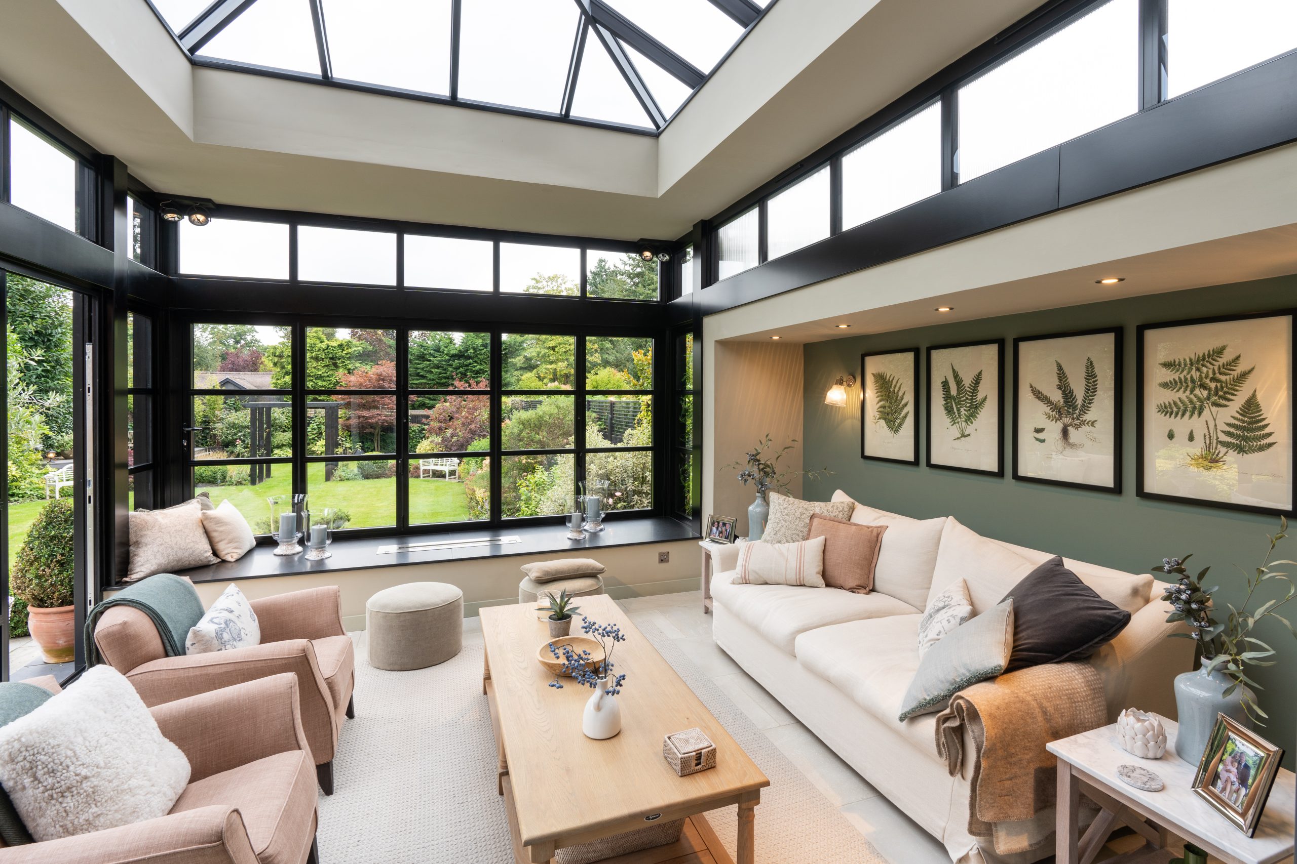 Interior living space within a black aluminium glazed extension, featuring roof glazing, large windows overlooking the garden, a neutral sofa seating area, and framed botanical artwork on the wall.